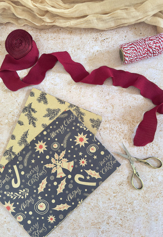 Overhead view of Christmas gift wrapping supplies including two sheets of patterned paper, a roll of burgundy ribbon, a spool of red and white twine, and a pair of ornate scissors on a textured surface.