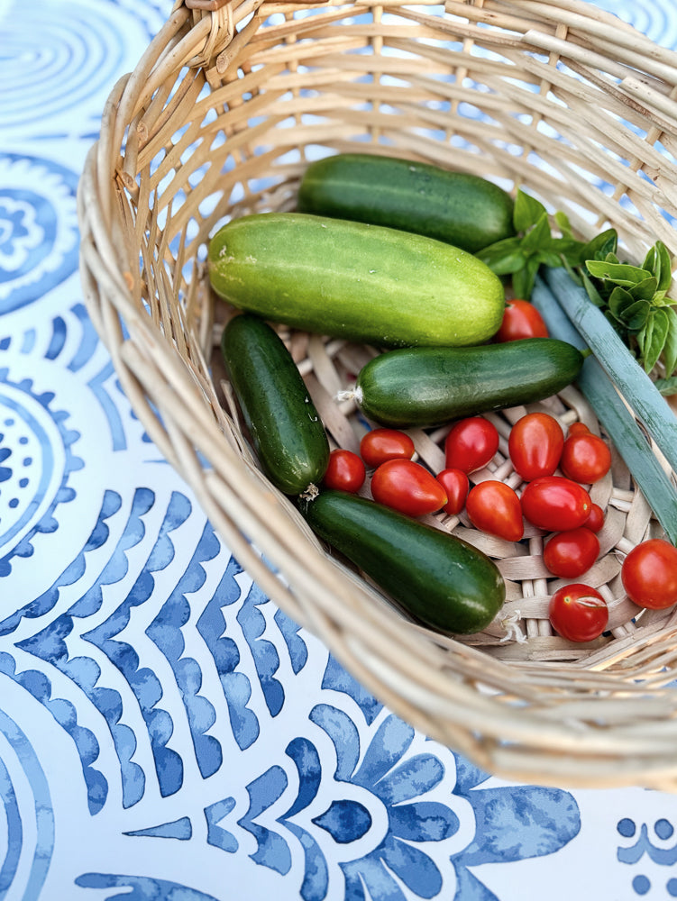 A woven basket filled with fresh vegetables, including cucumbers, cherry tomatoes, and green onions, rests on a blue and white patterned surface.