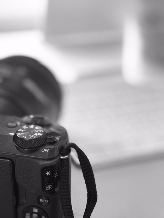 Close-up of the top of a black digital camera, showing the mode dial with settings like AV, TV, P, and OFF. A black camera strap is attached to the side.