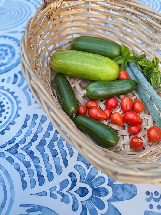 A woven basket filled with fresh vegetables, including cucumbers, cherry tomatoes, and green onions, rests on a blue and white patterned tablecloth.
