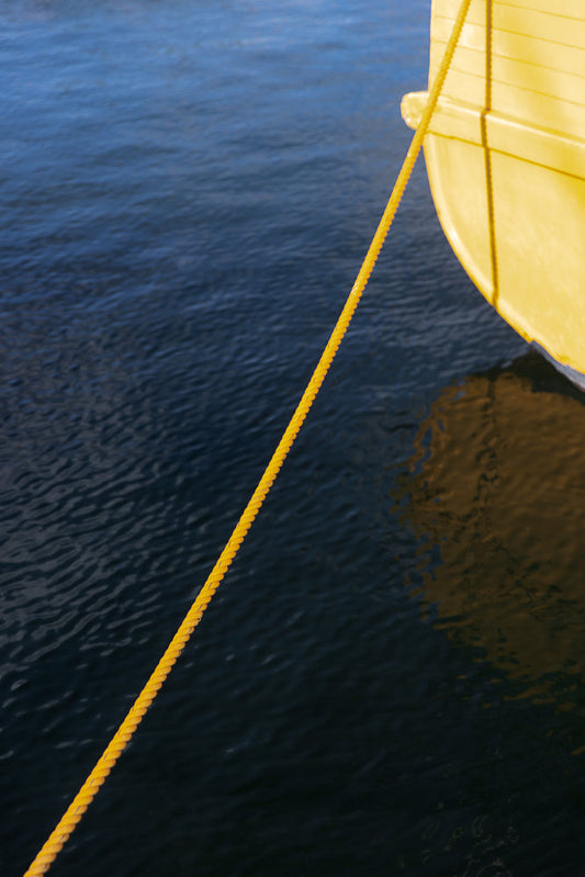 A bright yellow rope extends diagonally across the frame, connecting a yellow boat on the right to the dark blue water on the left. The rope is thick and textured, with a visible braid pattern. The water is calm with subtle ripples, reflecting the yellow of the boat in places.