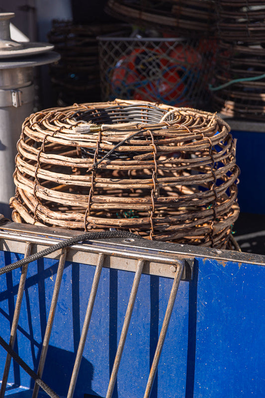 A close-up shot of a woven crab pot with a black rope draped over the top. The pot is made of natural materials and is secured with wire. In the background, other crab pots and fishing equipment are visible.