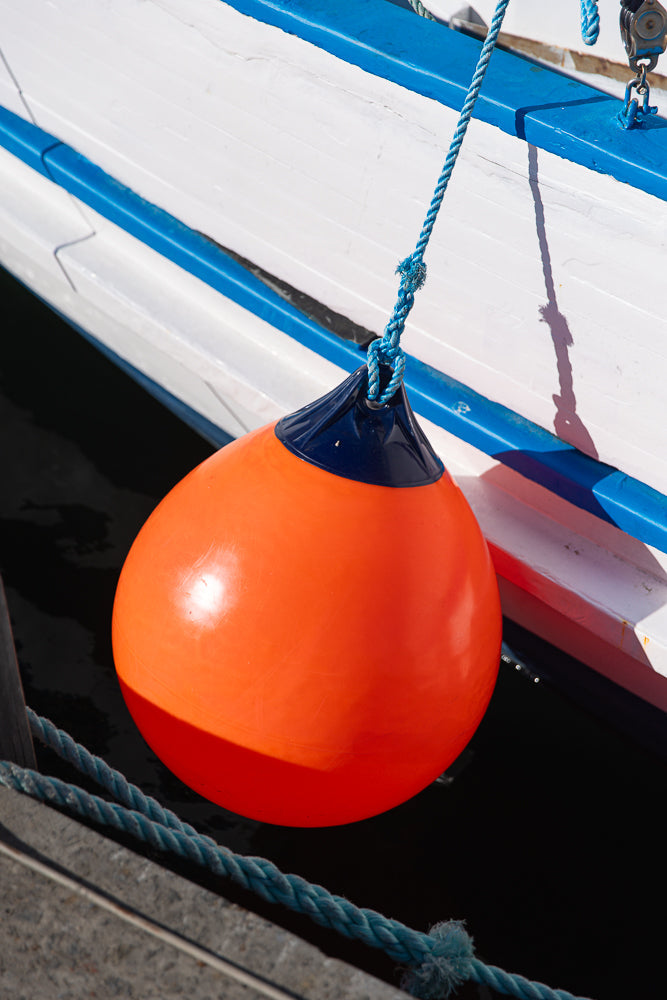 A bright orange buoy with a dark blue top hangs from a thick blue rope attached to a white boat with a blue stripe.