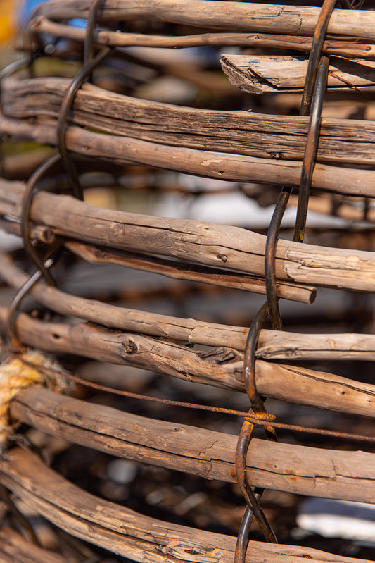 Close-up of a rustic woven basket made of natural wood branches, bound together with dark metal wire. The texture of the wood is rough and weathered, with visible grain and knots.