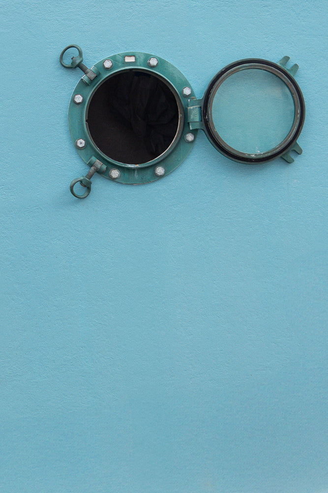 A round, teal-colored porthole window is mounted on a textured blue wall. The window is open, with the glass pane swung upwards and secured by a metal latch. The metal frame is bolted to the wall with visible bolts.