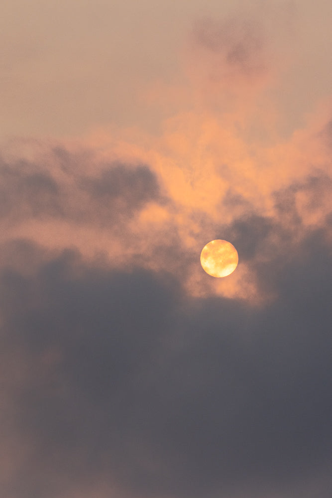 The sun is partially obscured by dark clouds, casting a warm orange glow through the sky. The clouds are a mix of dark gray and lighter, pinkish-orange hues, creating a dramatic and atmospheric scene.
