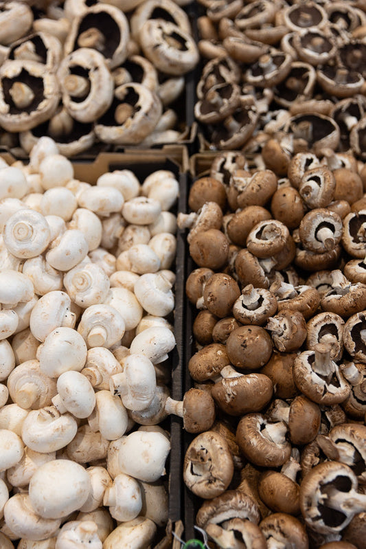 A close-up, overhead view shows two types of mushrooms in separate containers. On the left are white button mushrooms, and on the right are brown cremini mushrooms.