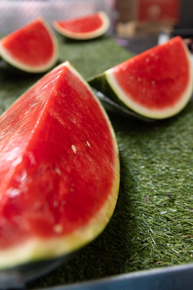 Several slices of watermelon are arranged on a green surface. The closest slice is in sharp focus, showing its vibrant red flesh and a few seeds. Other slices are visible in the background, slightly out of focus.