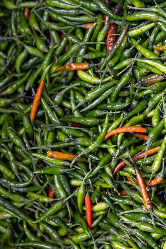A close-up, top-down view of a pile of fresh green chili peppers, with a few bright orange and red peppers interspersed throughout. The peppers are small and slender with pointed tips and green stems.