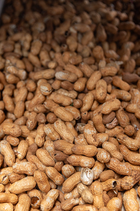 A close-up, overhead view of a large pile of unshelled peanuts. The peanuts are light brown with textured shells and are densely packed together.