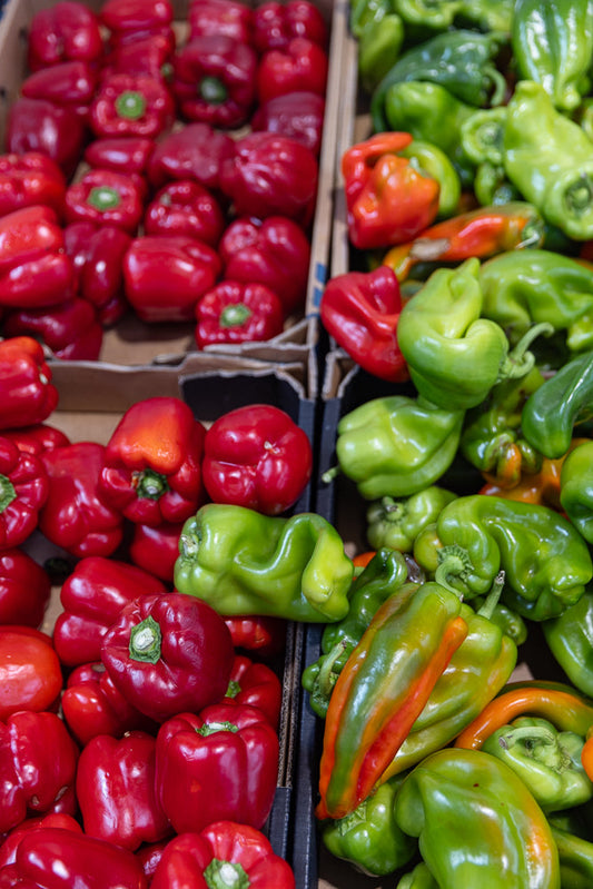 A close-up, high-angle shot shows a vibrant display of fresh bell peppers. On the left, a bin is filled with glossy, deep red bell peppers. To the right, another bin overflows with bright green bell peppers, some showing hints of orange and red, indicating ripening.