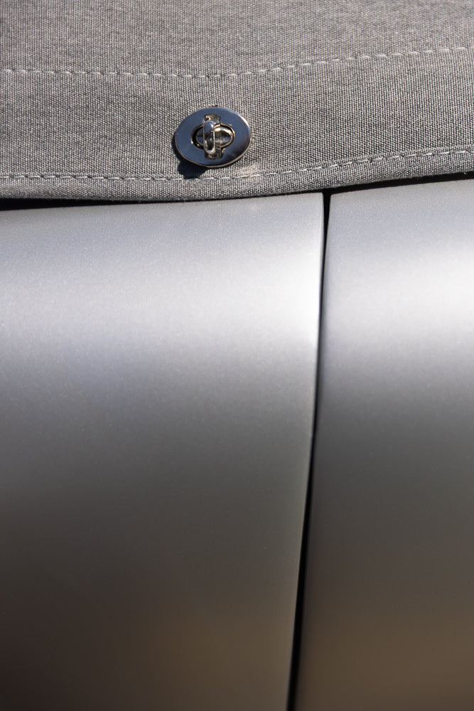 A close-up shot shows a silver metal clasp on a gray fabric surface, with a seam running horizontally above the clasp. The background is a smooth, reflective silver surface, possibly a car door or a metallic object.