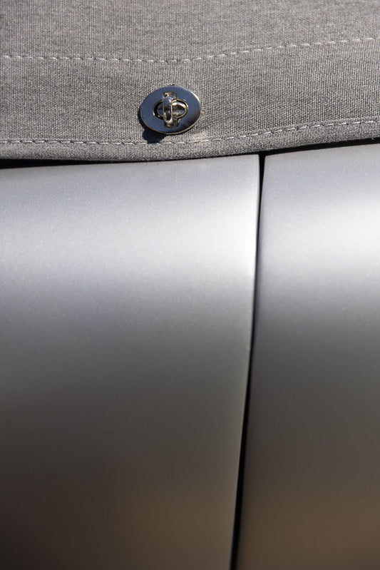 A close-up shot shows a silver metal clasp on a gray fabric surface, with a seam running horizontally above the clasp. The background is a smooth, reflective silver surface, possibly a car door or a metallic object.