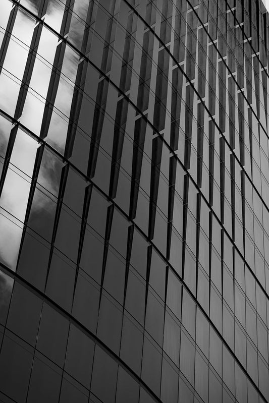 A black and white, low-angle shot of a modern building with a glass facade. The glass panels are arranged in a grid pattern, with some panels reflecting the sky and clouds. The building's architecture creates a sense of depth and repetition.