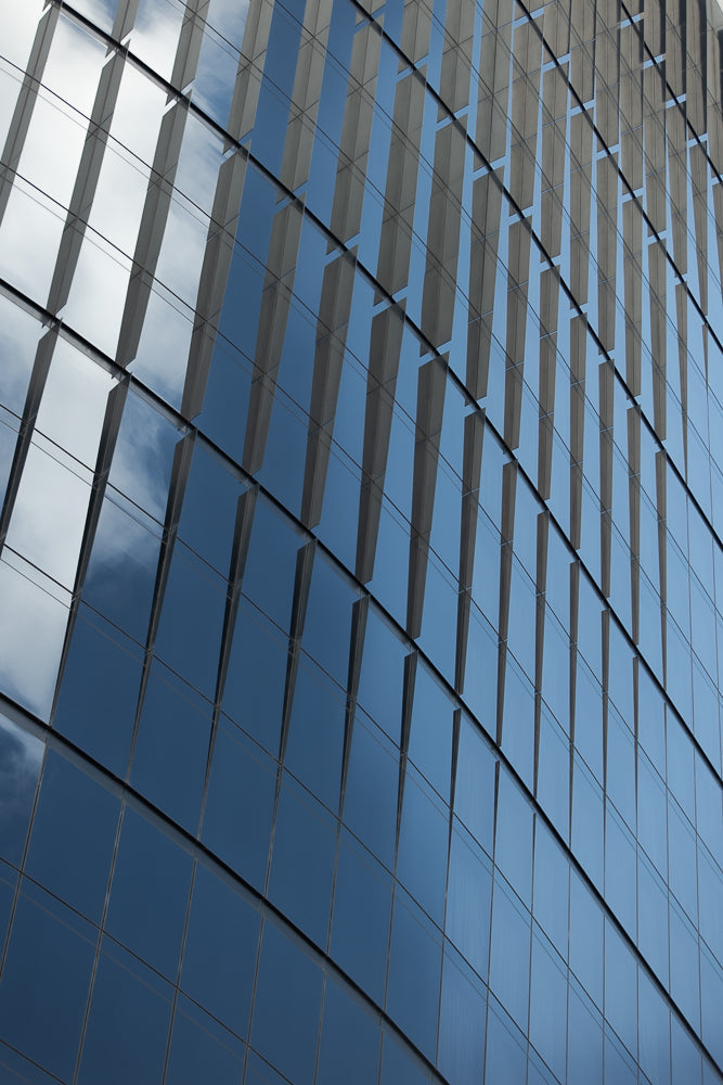 A modern building facade with a grid of reflective glass panels. The panels show a reflection of the sky with clouds and some darker blue sky areas. The building's structure creates a repeating pattern of vertical elements.