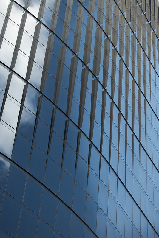 A modern building facade with a grid of reflective glass panels. The panels show a reflection of the sky with clouds and some darker blue sky areas. The building's structure creates a repeating pattern of vertical elements.