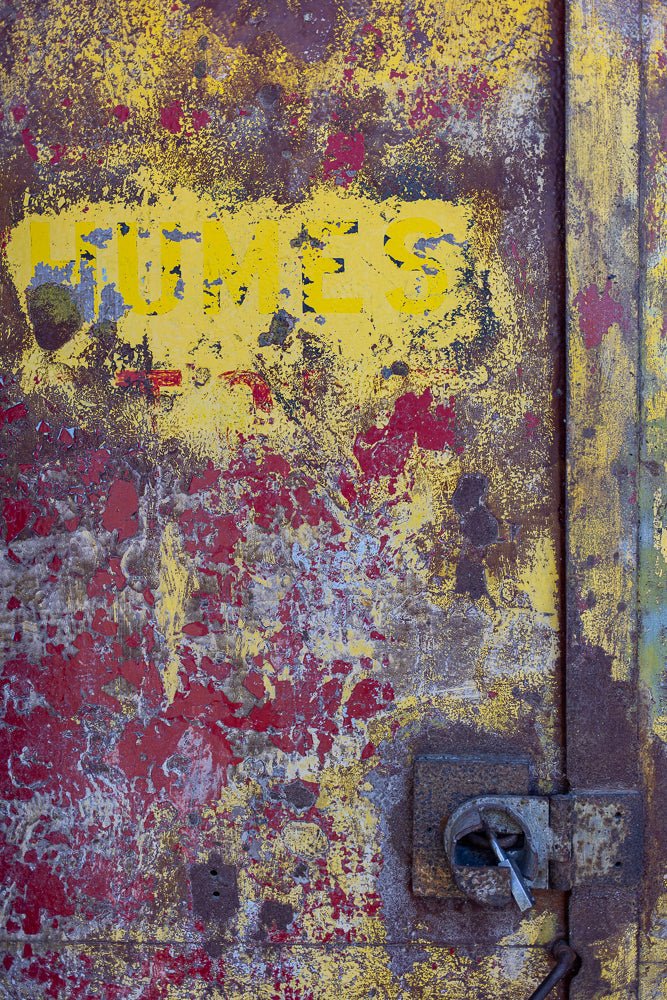 A close-up, abstract view of a heavily weathered and rusted metal surface. The surface is painted with peeling layers of yellow and red paint, revealing patches of dark brown rust and some lighter grey metal underneath. The word "HUMES" is visible in faded yellow block letters across the top portion of the image. A rusted metal lock and hasp are attached to the right side of the surface.