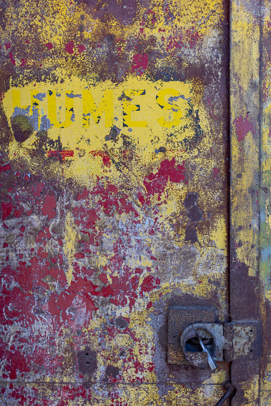 A close-up, abstract view of a heavily weathered and rusted metal surface. The surface is painted with peeling layers of yellow and red paint, revealing patches of dark brown rust and some lighter grey metal underneath. The word "HUMES" is visible in faded yellow block letters across the top portion of the image. A rusted metal lock and hasp are attached to the right side of the surface.