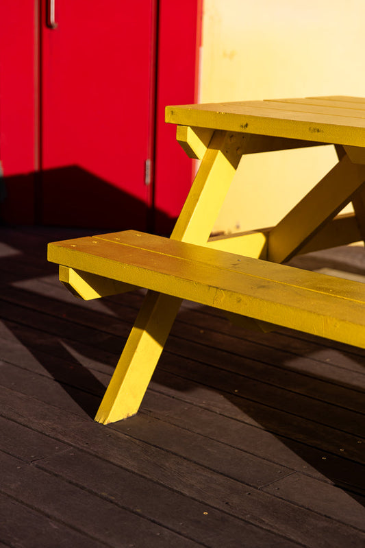 A bright yellow picnic table with a red door and yellow wall in the background. The table is casting a shadow on the dark wooden planks of the ground.