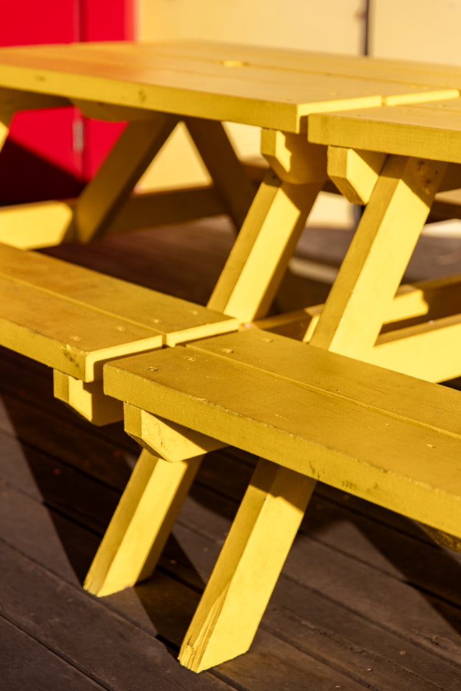 A bright yellow picnic table with attached benches sits on a dark wooden deck. The table and benches are made of wood and are painted a vibrant yellow. The sun casts shadows on the deck and the table legs.