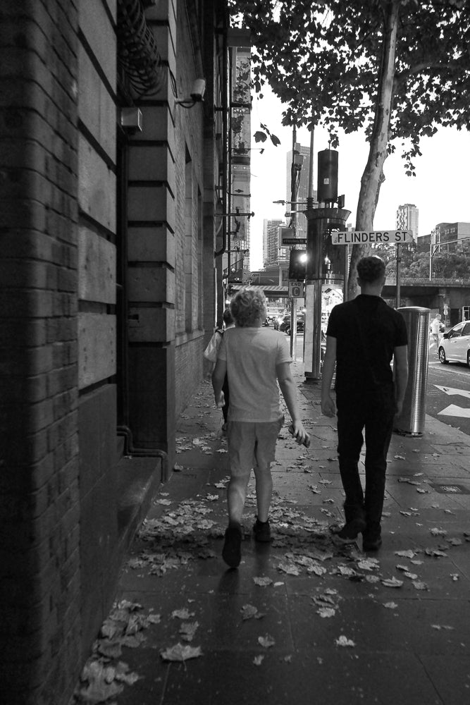 Two people walk down a sidewalk covered in fallen leaves. A street sign for Flinders St is visible in the background, along with city buildings and traffic.