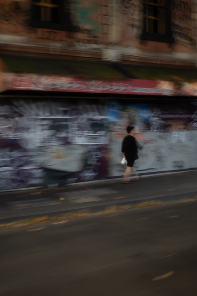 A person walks past a graffiti-covered wall. The image is blurred due to motion, giving a sense of movement and urban atmosphere.