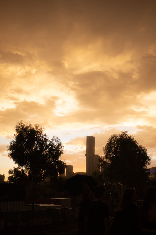 A dramatic sunset with golden clouds over a city skyline. Silhouettes of trees and people are visible in the foreground.