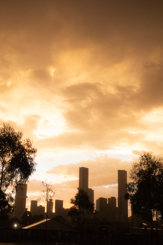 A city skyline is silhouetted against a dramatic orange and yellow cloudy sky at sunset. Trees in the foreground are also silhouetted, adding depth to the scene.