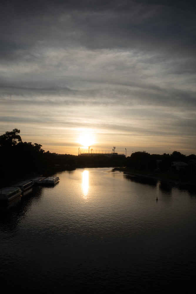 A wide shot of a river at sunset. The sun is low on the horizon, casting a bright reflection on the water. Several boats are docked on the left side of the river, and trees line the banks. A stadium with lights is visible in the distance.