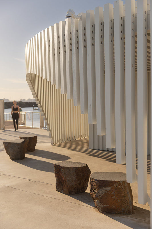 A woman in athletic wear walks past rough-hewn stone seats in front of a modern white slatted structure on a sunny day.
