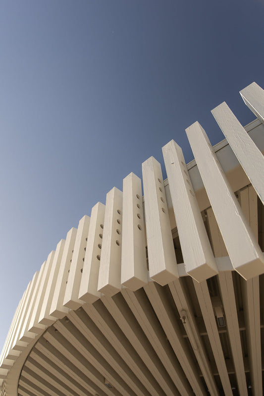 A low-angle shot of a white wooden structure against a clear blue sky. The structure is composed of vertical slats with circular holes, arranged in a curved pattern.