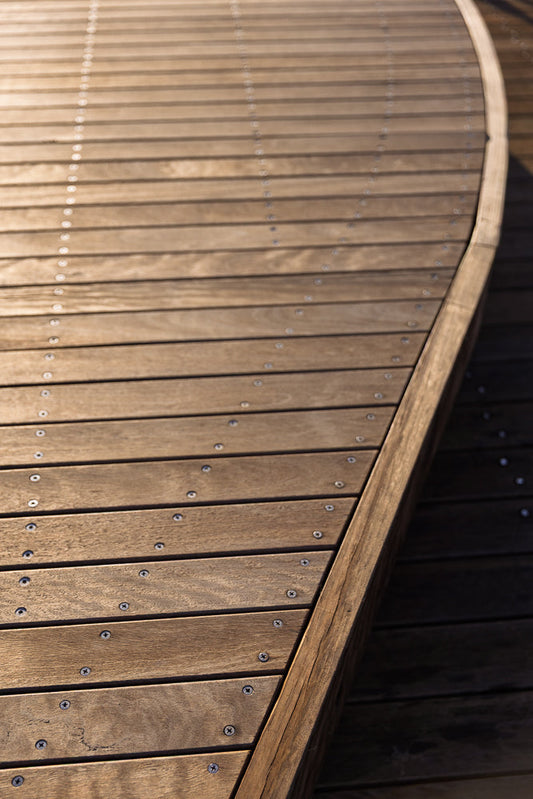 A close-up, angled view of a curved wooden deck made of horizontal planks. The planks are fastened with visible screws, and the wood shows a natural grain pattern. Sunlight casts shadows, highlighting the texture and the curve of the structure.