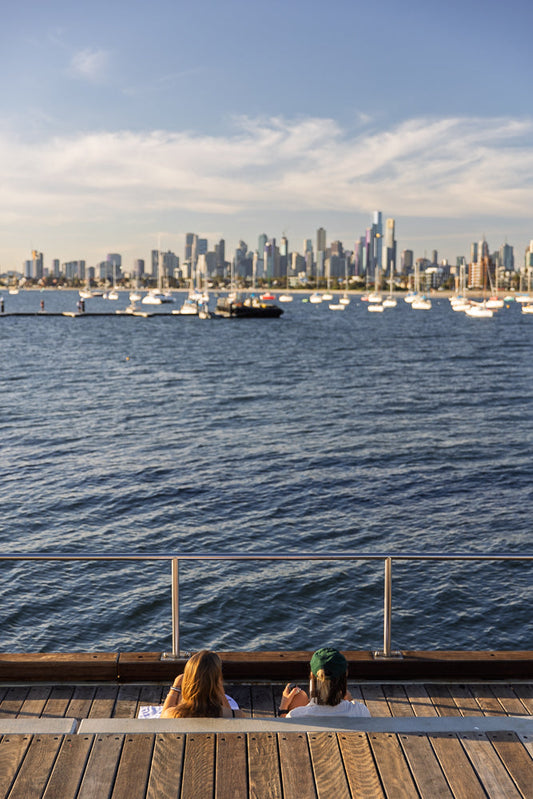 Two people sit on a wooden deck overlooking a bay filled with sailboats and a city skyline in the distance.