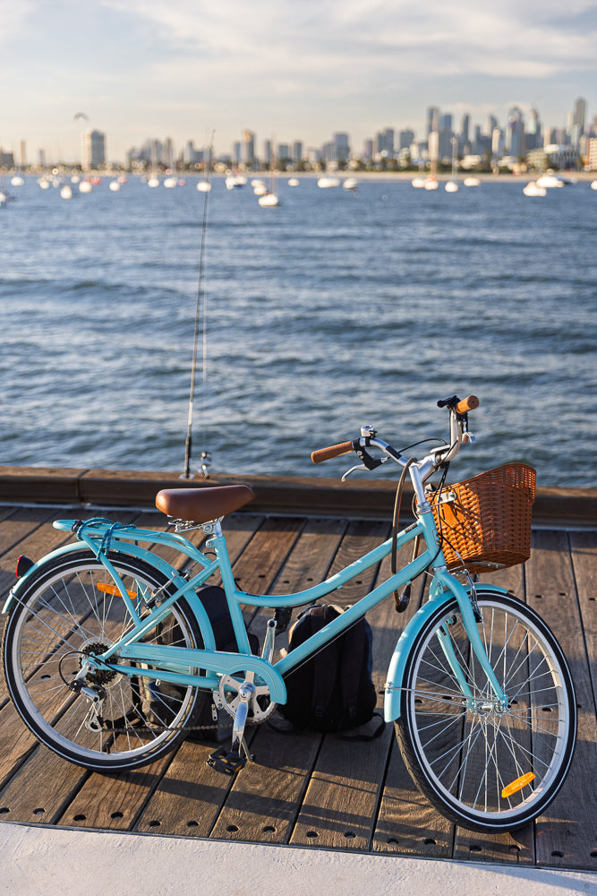 A light blue cruiser bicycle with a wicker basket is parked on a wooden pier. The ocean is in the background with sailboats and a city skyline.