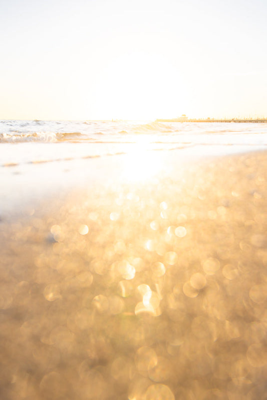 A low-angle shot of a beach with the sun reflecting off the wet sand and the ocean waves. The background shows a pier and a clear sky.