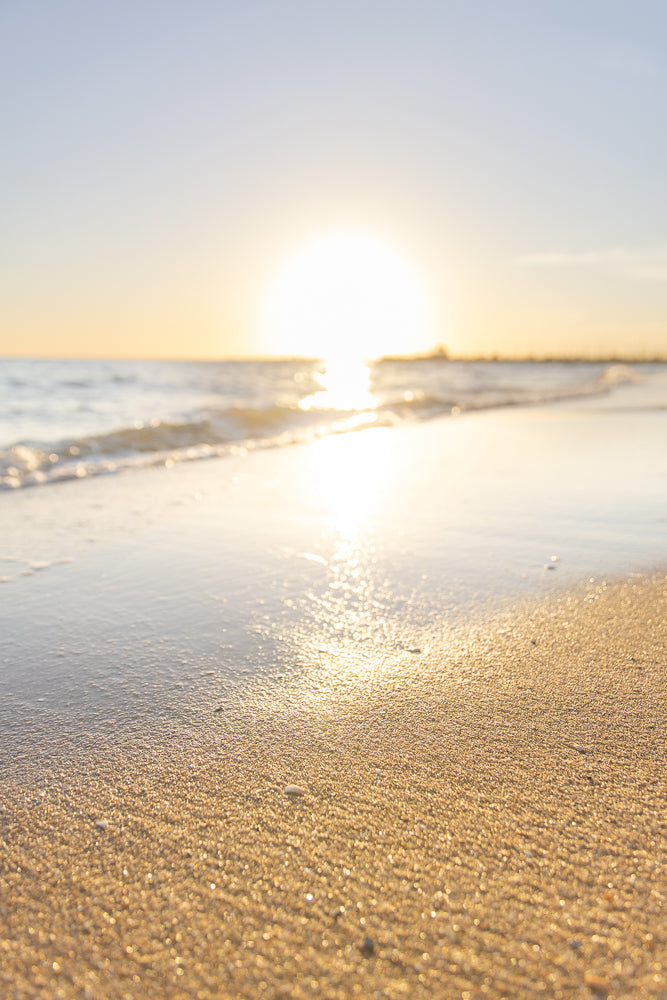 A low-angle shot of a sandy beach with gentle waves lapping at the shore during sunset. The sun is low on the horizon, casting a warm golden glow across the water and sand.