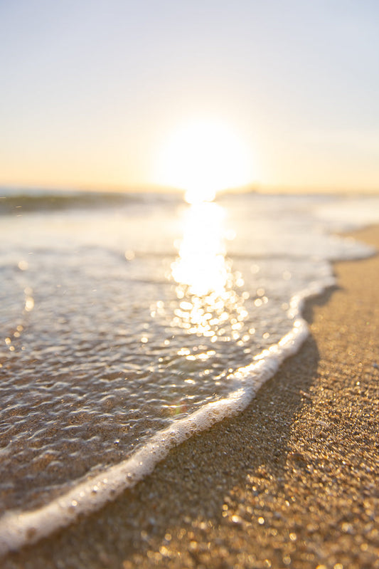 A gentle wave washes onto a sandy beach at sunset. The sun reflects brightly on the water, creating a shimmering path across the ocean. The sand is golden and sparkling, with small shells and pebbles visible.