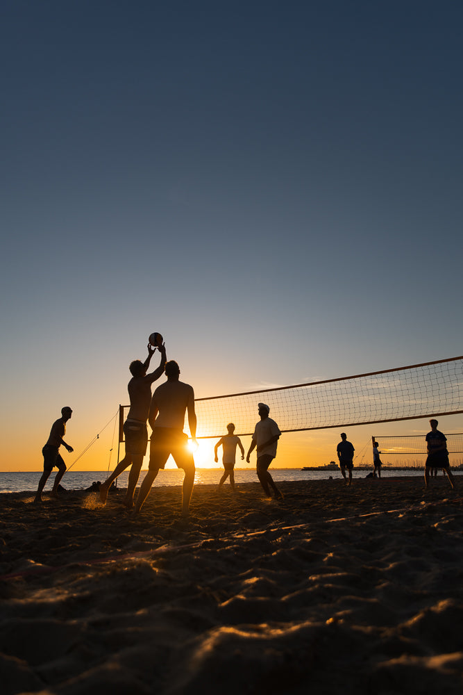 Silhouettes of people playing beach volleyball at sunset. The sun is low on the horizon, casting a warm glow and creating lens flare. The sky is a gradient of blue and orange.