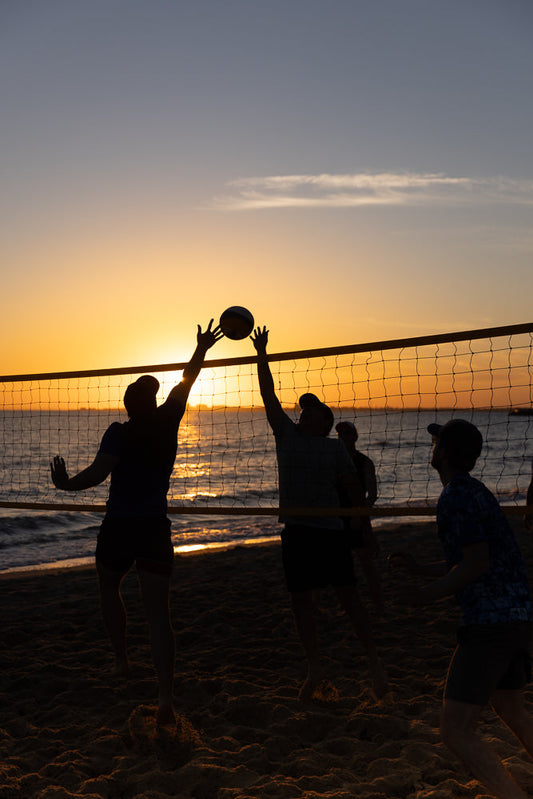 Silhouettes of people playing beach volleyball at sunset. Two players jump to hit the ball over the net, with the ocean and a golden sky in the background.