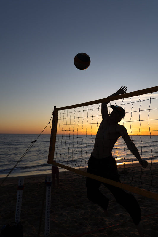 Silhouette of a beach volleyball player jumping to hit the ball over the net at sunset. The sky is a gradient of blue to orange, and the ocean is visible in the background.