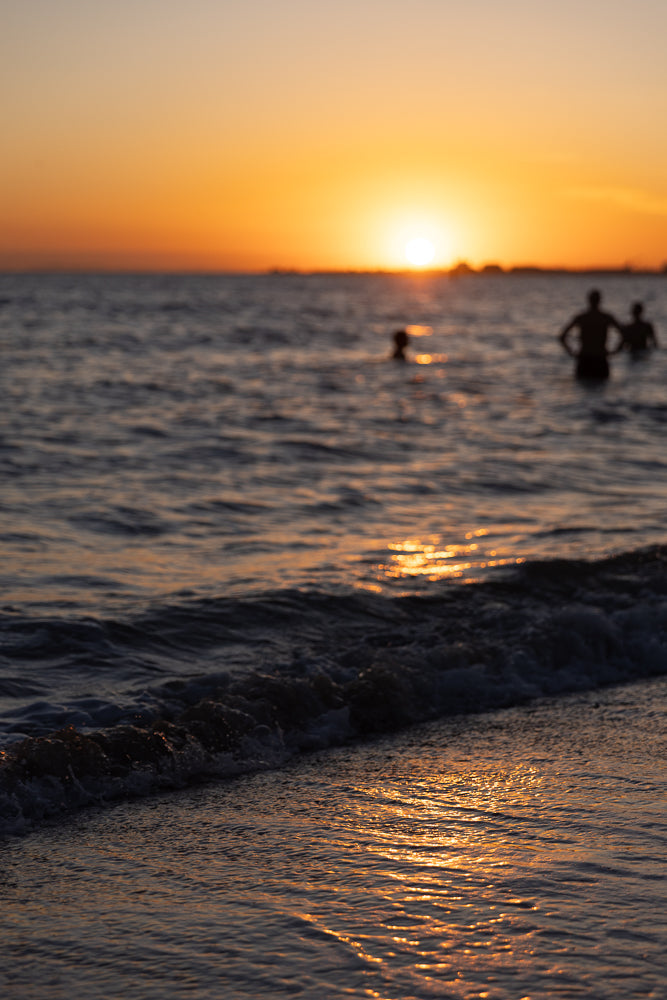 Silhouettes of people wading in the ocean at sunset. The sun is low on the horizon, casting a warm orange glow across the water and sky. Gentle waves lap at the shore in the foreground.