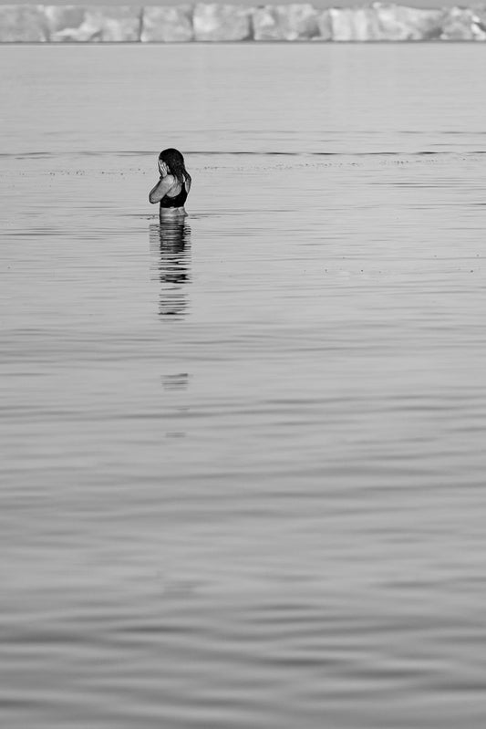 A person stands in calm water, with their hands covering their face. The background shows a blurred, rocky shoreline.