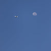 A small white airplane flies across a clear blue sky towards a large, pale moon.