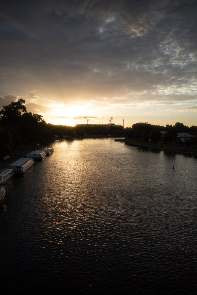 A wide river reflects the golden light of a setting sun. Several houseboats are moored along the left bank, and trees line the opposite bank. In the distance, a stadium with tall lights is silhouetted against the bright sky.