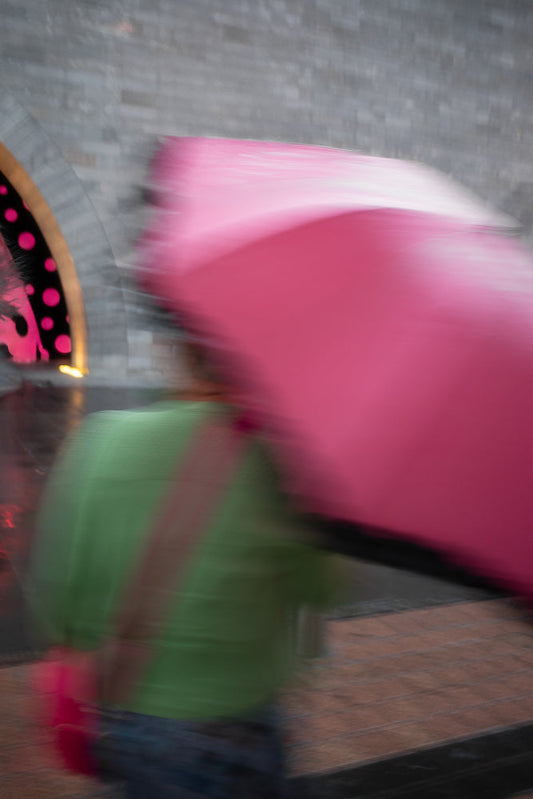 A person in a green shirt walks with a large pink umbrella. The image is blurred due to motion, giving a sense of movement.