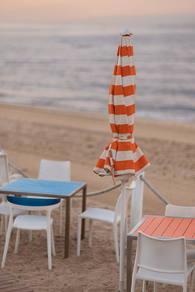 An orange and white striped umbrella is closed and tied with twine, standing on a sandy beach next to tables and chairs. The ocean is visible in the background under a soft, hazy sky.