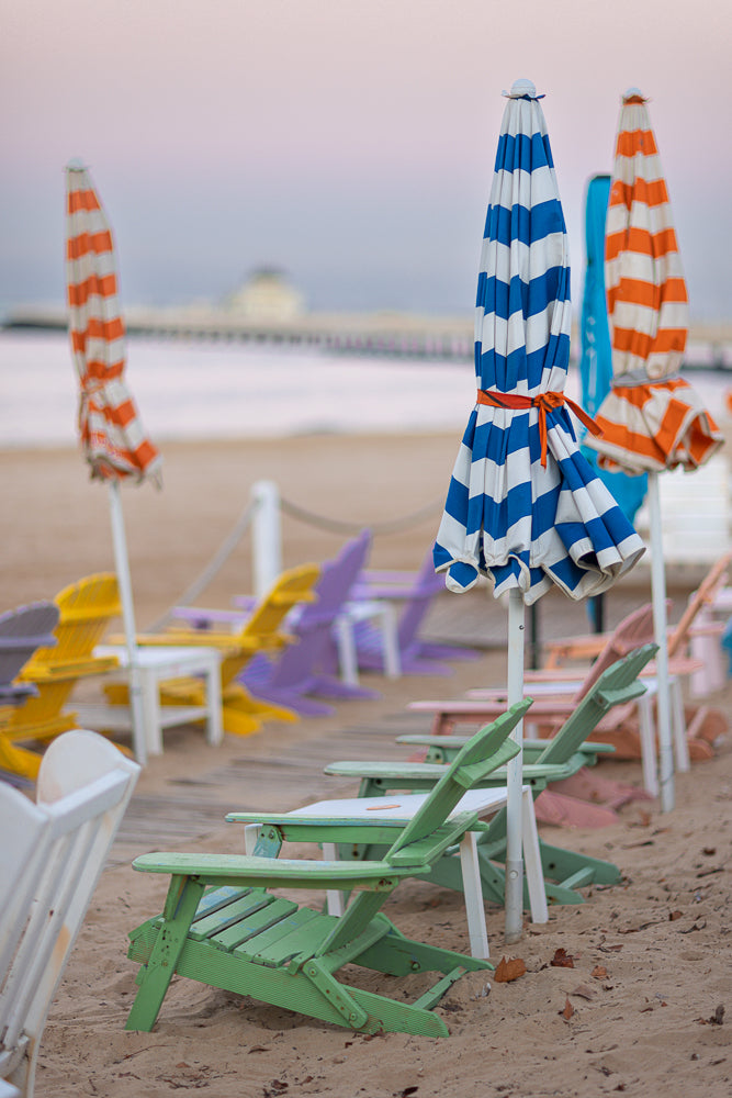 A row of colorful Adirondack chairs and closed beach umbrellas sit on a sandy beach at dusk. The sky is a soft pink and purple gradient.