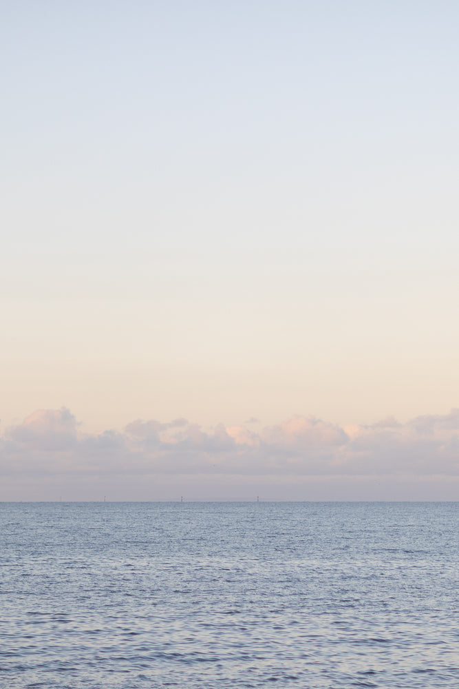 A serene seascape at dawn or dusk, with gentle waves on the water and soft, pastel-colored clouds on the horizon under a pale sky. Several distant buoys or markers are visible in the water.