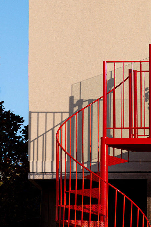 A bright red spiral staircase is shown against a beige wall and a blue sky. The staircase casts a shadow on the wall. Dark foliage is visible on the left side of the image.