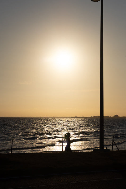 Silhouette of a person with a ponytail walking along the shore at sunset. The sun reflects off the choppy water, and a tall lamppost stands to the right.